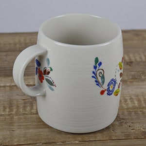 May include: Cream-colored ceramic mug with a curved handle and floral design. The flowers are in shades of red, blue, green, and brown. The mug sits on a wooden surface.