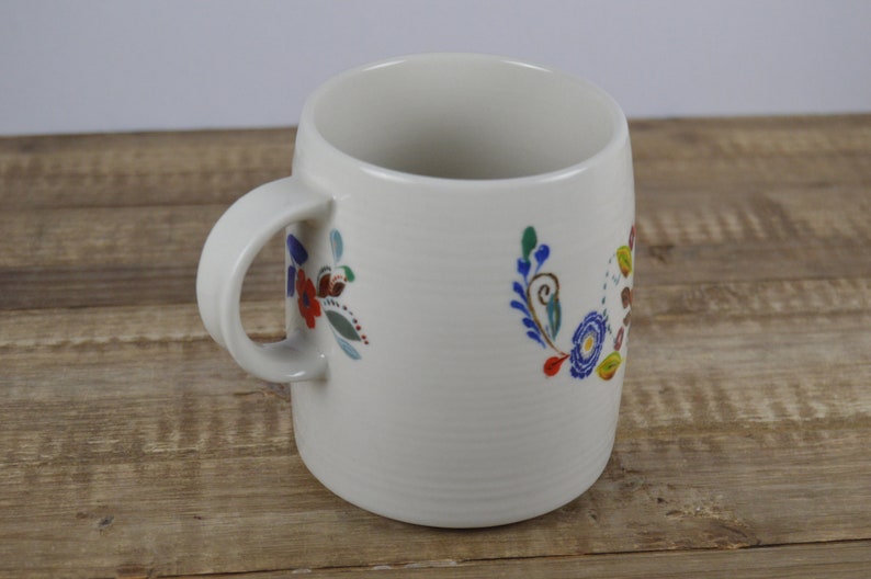 May include: Cream-colored ceramic mug with a curved handle and floral design. The flowers are in shades of red, blue, green, and brown. The mug sits on a wooden surface.