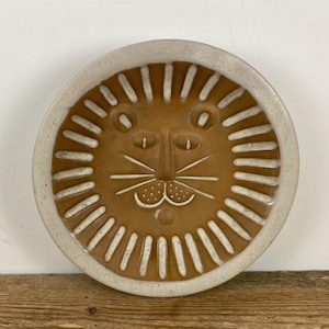 May include: A round ceramic plate featuring a stylized lion face in the center. The lion has a brown face with white incised lines for the mane, whiskers, eyes, and mouth. The plate has a wide, speckled off-white rim surrounding the lion.