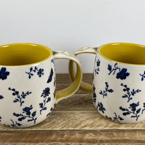 May include: Two ceramic mugs with a shared handle. The mugs have a white exterior with a blue floral pattern and a yellow interior. The mugs are on a wooden surface.