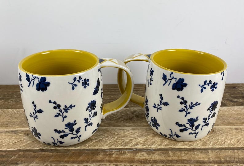 May include: Two ceramic mugs with a shared handle. The mugs have a white exterior with a blue floral pattern and a yellow interior. The mugs are on a wooden surface.