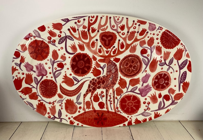 Op de afbeelding: Een ovale keramische schaal met een cr&egrave;mekleurige achtergrond, met een hert en bloemenontwerp in tinten rood en paars. Het hert heeft een geometrisch patroon en de bloemen zijn onder meer tulpen en gestileerde bloemen. De schaal is ongeveer 46 cm lang.