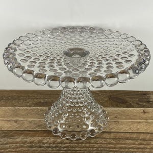 May include: A clear glass cake stand with a round top and a pedestal base. The top and base are decorated with a pattern of small, round, bubble-like details. The stand is designed to hold and display cakes or other desserts.