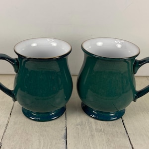 May include: Two matching ceramic mugs with a deep forest green exterior glaze and a smooth white interior. Each mug features a rounded, bulbous body, a sturdy curved handle on the side, and a raised pedestal base with a dark rim around the top edge.
