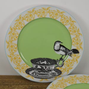 K&ouml;nnte beinhalten: Ein gr&uuml;ner und wei&szlig;er Teller aus Keramik mit einem gelben und goldenen Blumensaum. In der Mitte des Tellers befindet sich eine Schwarzwei&szlig;illustration eines Vogels, der auf einer Teetasse sitzt.