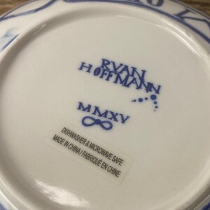 Puede incluir: Un plato de cer&aacute;mica blanco con ribete decorativo azul. La parte inferior del plato presenta el texto "RYAN HOFFMANN" y "MMXV" en azul. Una etiqueta dice "APTO PARA LAVAVAJILLAS Y MICROONDAS FABRICADO EN CHINA/FABRIQU&Eacute; EN CHINE".