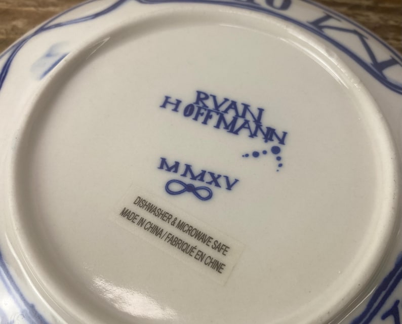 Puede incluir: Un plato de cer&aacute;mica blanco con ribete decorativo azul. La parte inferior del plato presenta el texto "RYAN HOFFMANN" y "MMXV" en azul. Una etiqueta dice "APTO PARA LAVAVAJILLAS Y MICROONDAS FABRICADO EN CHINA/FABRIQU&Eacute; EN CHINE".