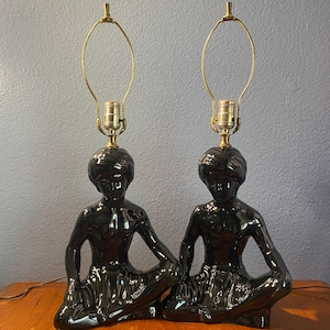 May include: Pair of black ceramic lamps featuring a seated figure with a glossy finish. Each lamp has a brass-colored harp and a bulb socket. The lamps are placed on a wooden surface against a blue wall.