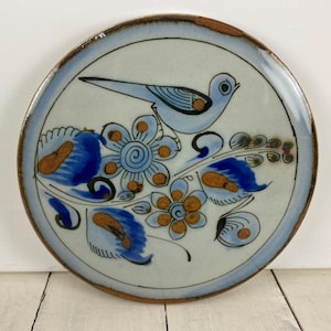 May include: Round ceramic plate with a hand-painted design. The plate features a light blue bird, blue flowers, and brown accents on a cream background. The rim is a darker brown with a light blue border.