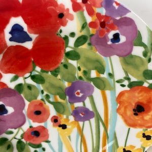 May include: A white ceramic plate with a vibrant floral design. The plate features large red, purple, and orange flowers with green leaves and blue and yellow accents. The flowers are painted in a watercolor style.