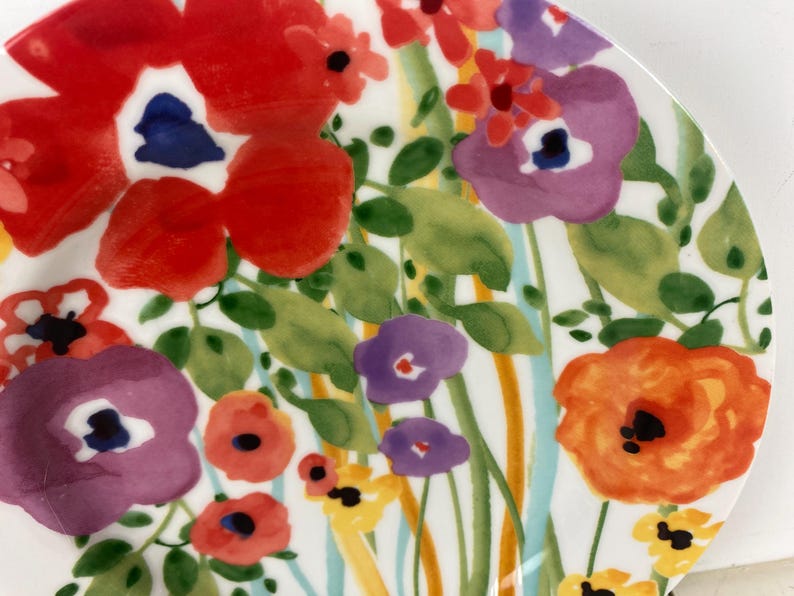 May include: A white ceramic plate with a vibrant floral design. The plate features large red, purple, and orange flowers with green leaves and blue and yellow accents. The flowers are painted in a watercolor style.