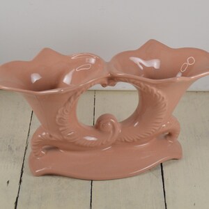 May include: A pink ceramic double vase with a unique design featuring two trumpet-shaped openings and a decorative base.