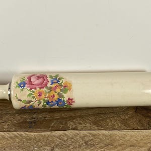 May include: Cream-colored rolling pin with a floral cross-stitch design. The pin has a cylindrical body with rounded handles and a loop on one end. The cross-stitch features pink roses, blue and yellow flowers.