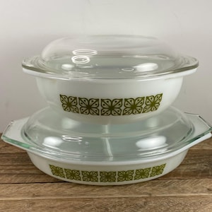 May include: Two white glass casserole dishes with clear glass lids. The dishes feature a repeating olive green floral pattern around the exterior. The top dish is round, while the bottom dish is oval-shaped with integrated side handles.