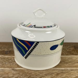 May include: White ceramic sugar bowl with lid. The bowl features a geometric design with blue, yellow, and red accents. The lid has a small handle and a similar design. The bowl is decorated with a dotted pattern and a spoon design.