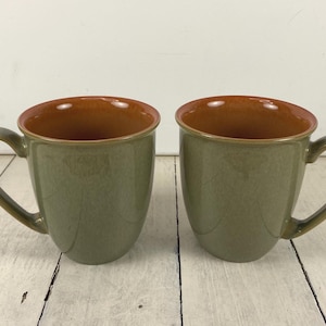 May include: Two ceramic mugs with a sage green exterior and a warm brown interior. The mugs have curved handles and sit on a white wooden surface. The mugs are designed for hot beverages.