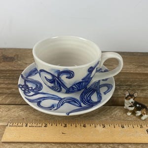 Puede incluir: Juego de taza y platillo de cer&aacute;mica blanca con dise&ntilde;os florales abstractos azules. La taza de t&eacute; tiene un asa y se apoya en un platillo a juego. Una peque&ntilde;a figura de corgi est&aacute; al lado de la taza y el platillo.