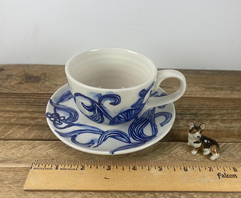 Puede incluir: Juego de taza y platillo de cer&aacute;mica blanca con dise&ntilde;os florales abstractos azules. La taza de t&eacute; tiene un asa y se apoya en un platillo a juego. Una peque&ntilde;a figura de corgi est&aacute; al lado de la taza y el platillo.