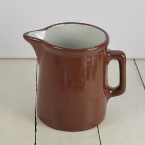 May include: A brown ceramic creamer with a white interior. The creamer has a rounded body and a handle.