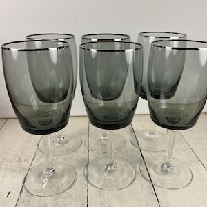 May include: Six gray wine glasses with a silver rim. The glasses have a clear stem and base. The glasses are arranged on a light-colored wooden surface.