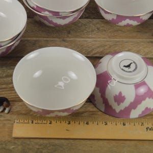 May include: Set of pink and white patterned ceramic bowls with gold trim. The bowls are stacked and arranged on a wooden surface. A small dog figurine is in the foreground.
