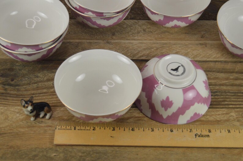May include: Set of pink and white patterned ceramic bowls with gold trim. The bowls are stacked and arranged on a wooden surface. A small dog figurine is in the foreground.