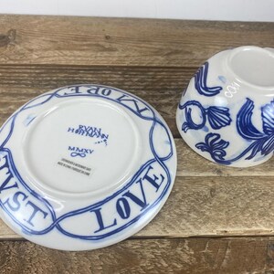 Puede incluir: Un juego de taza y platillo de cer&aacute;mica blanca con dise&ntilde;os florales azules y las palabras "JUST LOVE" en una pancarta azul. El platillo tiene el texto "RYAN HOFFMANN" y "MMXV".