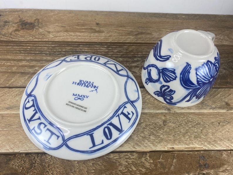 Puede incluir: Un juego de taza y platillo de cer&aacute;mica blanca con dise&ntilde;os florales azules y las palabras "JUST LOVE" en una pancarta azul. El platillo tiene el texto "RYAN HOFFMANN" y "MMXV".