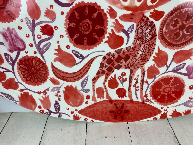 Op de afbeelding: Een witte keramische schaal met een rood en bordeaux bloemen- en dierenontwerp. Het ontwerp omvat een gestileerd hert met gedetailleerde patronen, bloemen en bladeren. De schaal heeft een afgeronde vorm.