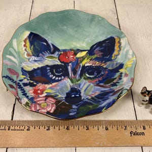 May include: A decorative ceramic plate with a raccoon face design. The plate has a scalloped edge and a gold rim. The raccoon is painted in shades of blue, green, and yellow, with red and pink floral accents. A small corgi figurine is next to the plate.