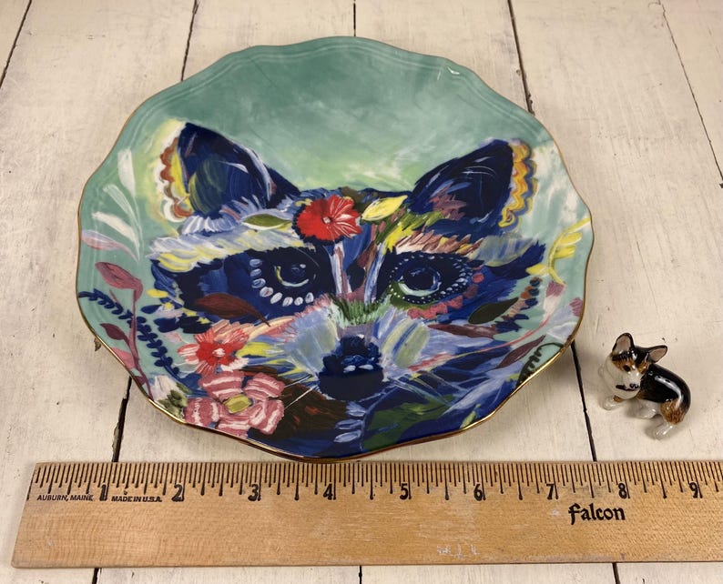 May include: A decorative ceramic plate with a raccoon face design. The plate has a scalloped edge and a gold rim. The raccoon is painted in shades of blue, green, and yellow, with red and pink floral accents. A small corgi figurine is next to the plate.