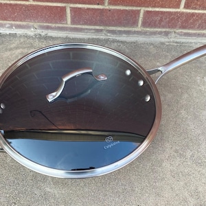 May include: Stainless steel frying pan with a glass lid. The pan has a long handle and a smaller loop handle on the side. The lid has a curved handle. The brand name "Calphalon" is visible on the pan.