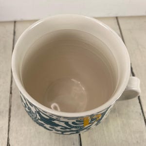 K&ouml;nnte beinhalten: Wei&szlig;er Keramikbecher mit einem t&uuml;rkis-wei&szlig;en Muster am Boden. Der Becher hat eine wei&szlig;e Innenseite und einen kleinen Henkel. Der Becher steht auf einer wei&szlig;en Holzoberfl&auml;che.
