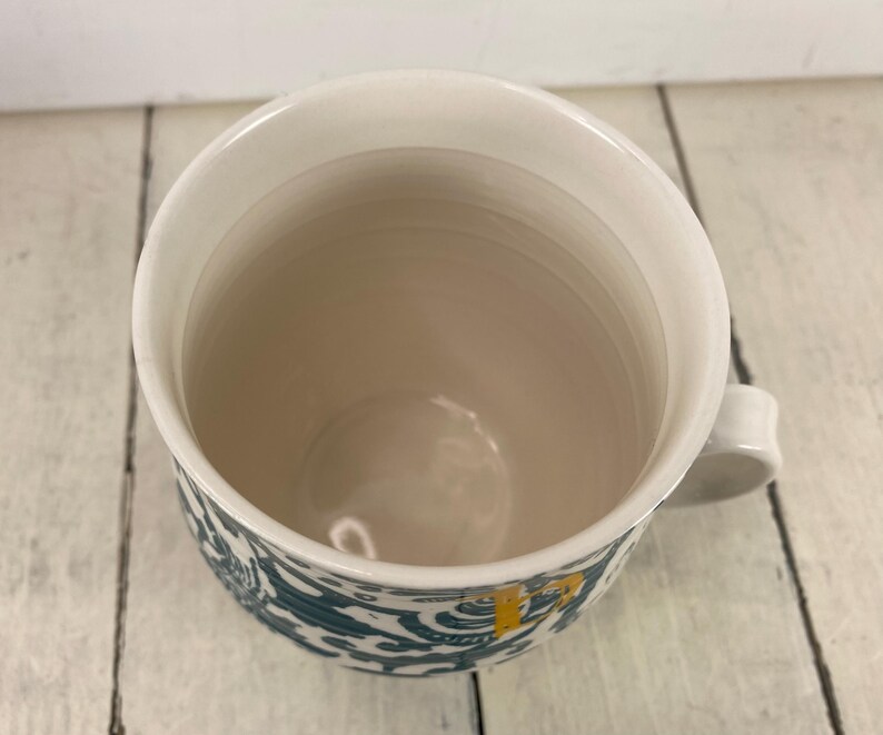 K&ouml;nnte beinhalten: Wei&szlig;er Keramikbecher mit einem t&uuml;rkis-wei&szlig;en Muster am Boden. Der Becher hat eine wei&szlig;e Innenseite und einen kleinen Henkel. Der Becher steht auf einer wei&szlig;en Holzoberfl&auml;che.