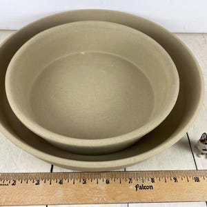 May include: Two beige stoneware bowls, one nested inside the other, with a small ceramic dog figurine to the side. A wooden ruler is in the foreground. The bowls have a simple, round design.