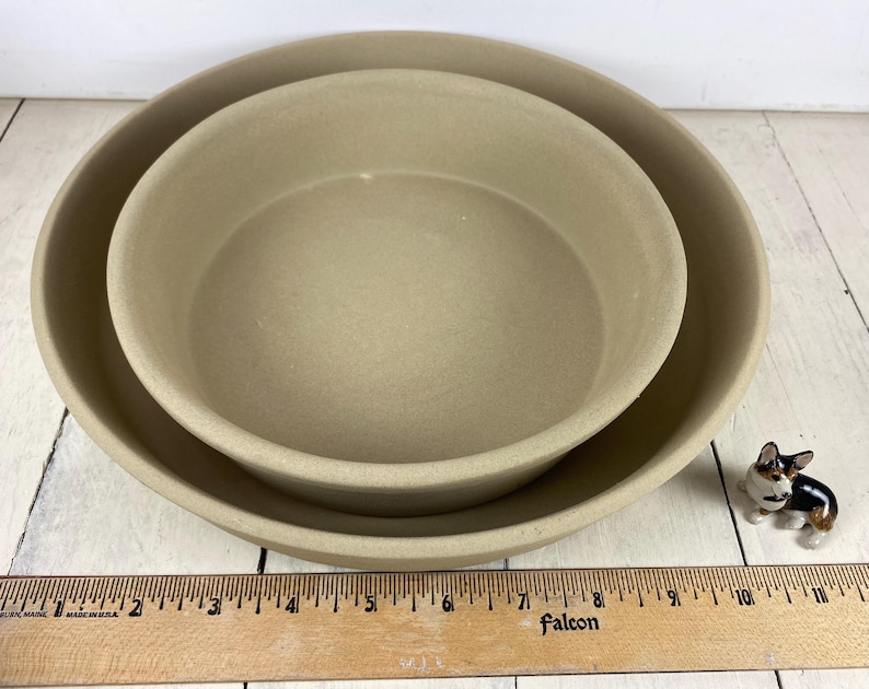 May include: Two beige stoneware bowls, one nested inside the other, with a small ceramic dog figurine to the side. A wooden ruler is in the foreground. The bowls have a simple, round design.