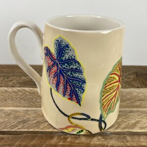 K&ouml;nnte beinhalten: Eine beige Keramiktasse mit gebogenem Henkel. Die Tasse hat bunte Blattmuster in Blau, Rot, Gelb und Gr&uuml;n. Die Tasse steht auf einer Holzoberfl&auml;che.