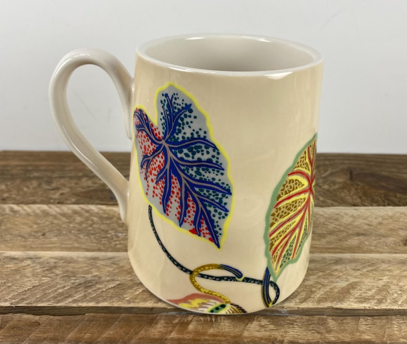 K&ouml;nnte beinhalten: Eine beige Keramiktasse mit gebogenem Henkel. Die Tasse hat bunte Blattmuster in Blau, Rot, Gelb und Gr&uuml;n. Die Tasse steht auf einer Holzoberfl&auml;che.
