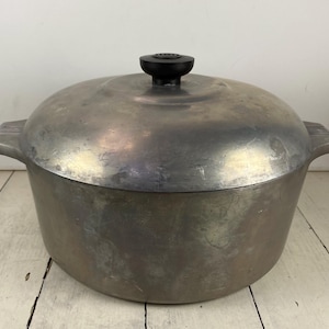 May include: A round, silver-colored metal Dutch oven pot with a matching lid. The pot features two integrated side handles with ribbed detailing and a small, circular black knob centered on the top of the lid. The metal surface shows signs of wear.