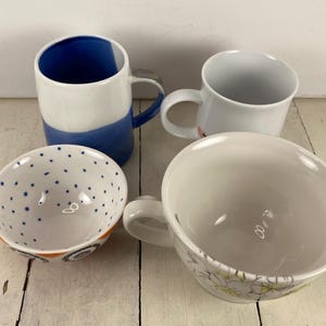 K&ouml;nnte beinhalten: Vier Keramikbecher und Schalen sind auf einer wei&szlig;en Holzoberfl&auml;che angeordnet. Ein Becher ist wei&szlig; mit einem blauen Abschnitt, ein anderer ist schlicht wei&szlig;, und ein dritter ist eine gro&szlig;e wei&szlig;e Schale mit einem Blumendesign. Eine kleine Schale hat blaue Punkte.