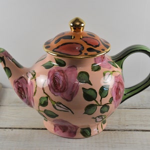 May include: A pink ceramic teapot with a green handle and lid. The teapot is decorated with a floral pattern of pink roses and green leaves. The lid has a leopard print design with gold accents.