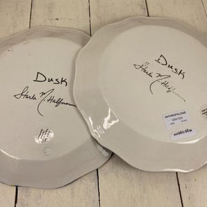 May include: Two white ceramic plates with scalloped edges. Each plate has the word "Dusk" and a signature in black ink. One plate has a sticker that says "Anthropologie". The plates are on a light wood surface.