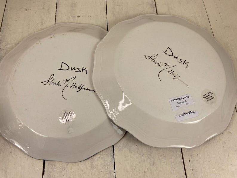 May include: Two white ceramic plates with scalloped edges. Each plate has the word "Dusk" and a signature in black ink. One plate has a sticker that says "Anthropologie". The plates are on a light wood surface.