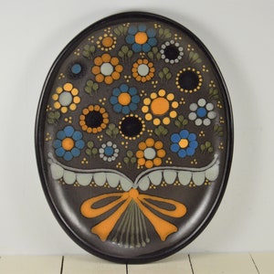 May include: A brown ceramic plate with a floral design. The flowers are yellow, blue, and white with black centers. The plate has a brown rim and a yellow bow at the bottom.