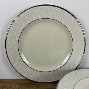 May include: A set of ivory-colored plates with a silver rim and a delicate, white floral pattern. The plates have a classic design, suitable for formal dining or special occasions. The plates are round and appear to be made of porcelain.
