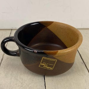 May include: A handcrafted stoneware soup bowl with a black handle and a color-blocked design in shades of brown and tan. The bowl has a label that reads "Pottery Craft".