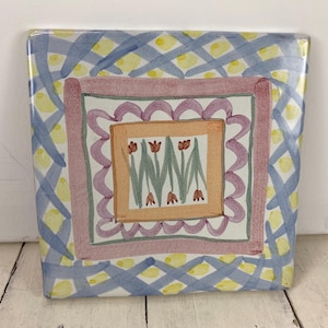May include: Square ceramic tile with a hand-painted design. The tile features a blue and yellow lattice pattern, a pink and purple border, and a central square with tulips. The tile is a decorative piece.
