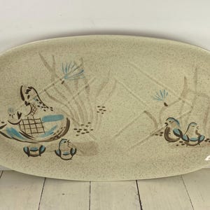 May include: Oval serving platter with a speckled beige surface. The platter features a hand-painted design of a duck with ducklings, and stylized aquatic plants in shades of blue, brown, and beige. The platter is a decorative piece.