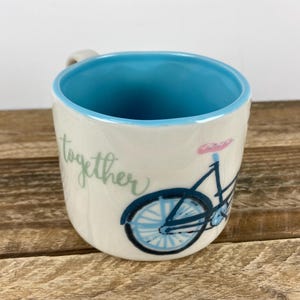 K&ouml;nnte beinhalten: Cremefarbene Keramiktasse mit hellblauem Innenraum und einer Fahrradillustration. Das Fahrrad ist schwarz und blau mit einem rosa Sattel. Das Wort "together" steht in Schreibschrift auf der Seite der Tasse.