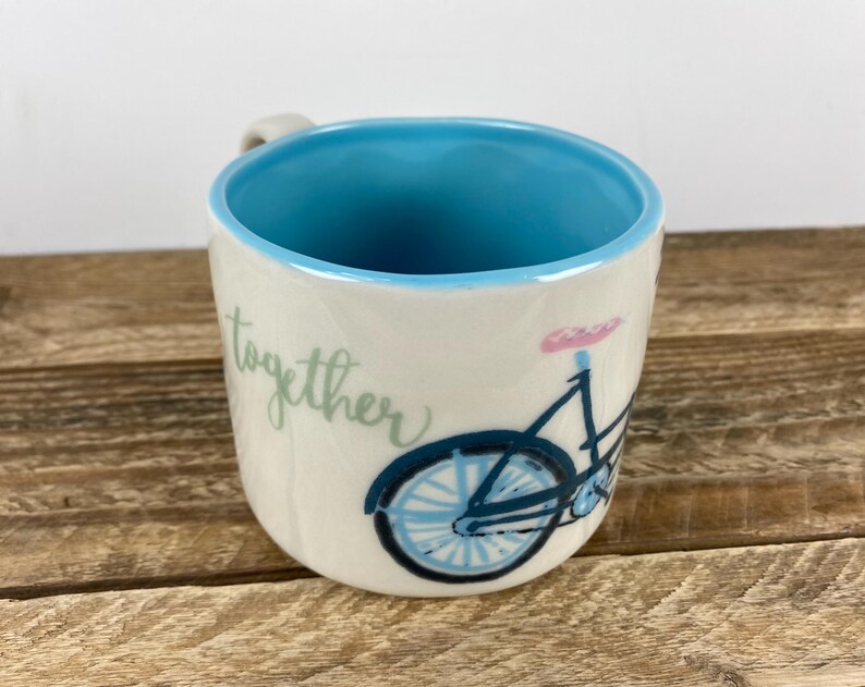 K&ouml;nnte beinhalten: Cremefarbene Keramiktasse mit hellblauem Innenraum und einer Fahrradillustration. Das Fahrrad ist schwarz und blau mit einem rosa Sattel. Das Wort "together" steht in Schreibschrift auf der Seite der Tasse.